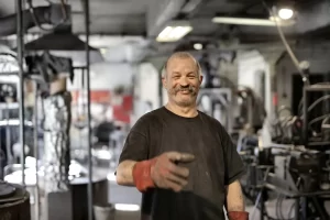 A smiling man wearing a black shirt and red work gloves stands in an industrial workshop, extending his hand toward the camera, as if offering a handshake. The background is filled with machinery and tools.