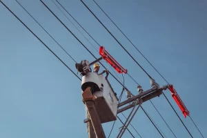 A worker in a hard hat stands in a raised bucket lift, inspecting or repairing overhead power lines against a clear blue sky. Some insulators and equipment on the lines are highlighted in red.