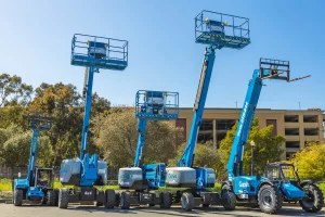 Several blue Genie construction lifts and boom lifts of various sizes are parked outdoors on pavement, surrounded by trees and a multi-level parking structure in the background.