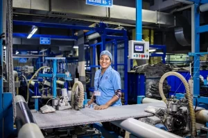 A woman wearing a blue uniform and hairnet smiles while working at a textile machine, surrounded by advanced manufacturing equipment in a brightly lit factory with blue machinery in the background.