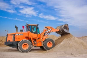 An orange Doosan DL300A wheel loader scoops a pile of sand at a construction site under a blue sky with scattered clouds, showcasing reliable performance that's ideal for projects considering Bulldozer Finance options.