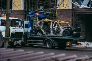 A used tow truck carries the charred remains of a burnt-out car on its flatbed in an urban area, with damaged buildings and bare trees visible in the background.