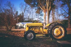 A yellow tractor, ideal for Landscape Equipment financing, is parked on fallen leaves near bare trees, with a white house and wooden fence in the background beneath a clear blue sky.