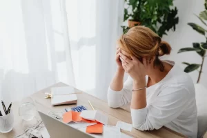 A woman sits at a desk with her head in her hands, looking stressed. Papers, receipts, money, and a laptop are spread out in front of her—she’s searching for ways to conserve cashflow. There are plants and sunlight in the background.