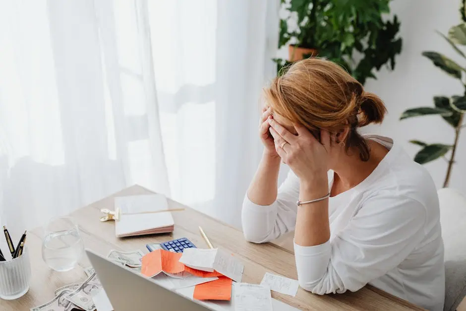 A woman sits at a desk with her head in her hands, looking stressed. Papers, receipts, money, and a laptop are spread out in front of her—she’s searching for ways to conserve cashflow. There are plants and sunlight in the background.