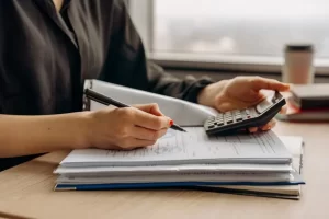 A business owner in a black shirt uses a calculator and writes on documents stacked on a desk, with an open binder beside them and a coffee cup in the background.