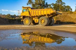 A large yellow dump truck, ideal for Truck Financing for Startups, is parked on a dirt road with its reflection visible in a puddle. Trees and a blue sky complete the scenic background.