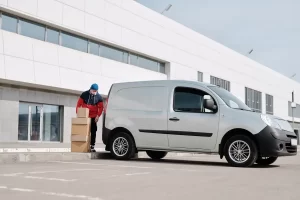 A delivery worker in a red and blue uniform and face mask unloads cardboard boxes from a white van—secured through Delivery Truck Financing—parked outside a modern, white commercial building.