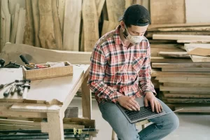 A man wearing a plaid shirt, jeans, backward cap, and face mask sits on a stool in a woodworking shop, using a laptop to manage his small business. Tools and wooden planks are visible around him.