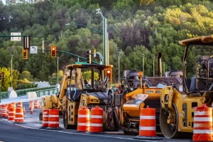 Several yellow construction vehicles and orange traffic barrels block a road under maintenance, with trees and traffic lights in the background—perfectly illustrating the need for affordable dump truck financing for large projects.