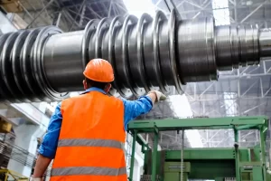 A worker wearing an orange safety vest and helmet stands in a factory, pointing towards a large CNC machine part suspended overhead in a spacious, well-lit facility.