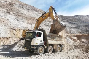 An excavator loads soil or rocks into a massive Dump Truck at a mining or construction site, set amid rocky, dusty terrain under a clear blue sky.