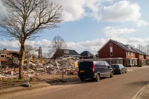 A fenced-off construction site with piles of debris and rubble from a demolished building sits beside a residential street lined with houses, parked cars, and a leafless tree—Backhoe Finance banners flutter under a partly cloudy sky.