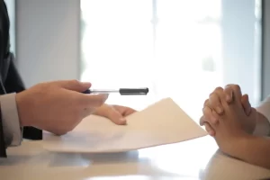 A person in business attire hands a pen and document to another, likely finalizing a Business Funding agreement during a meeting at a desk.
