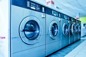 A row of large, stainless steel washing machines in a brightly lit laundromat, with tiled floors and a ceiling of fluorescent lights—an ideal setting for those considering financing laundry equipment.