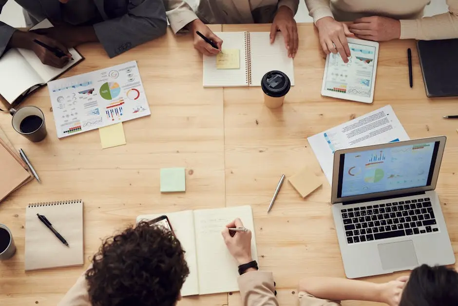 Four people sit around a wooden table with laptops, notebooks, charts, coffee cups, and papers, collaborating, taking notes, and reviewing documents during a business meeting.