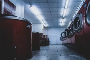 A low-angle view of an empty laundromat with red washing machines and dryers lining both sides, tiled floor, fluorescent ceiling lights, and a bench in the background.