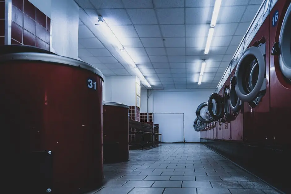 A low-angle view of an empty laundromat with red washing machines and dryers lining both sides, tiled floor, fluorescent ceiling lights, and a bench in the background.