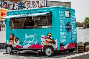 A bright turquoise ice cream truck with “Happy Beginning” written on it, decorated with images of ice cream treats and a menu listing items like kulfi, gelato, sundaes, and shakes. The truck is parked on a paved area.
