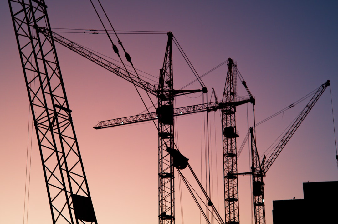 Silhouettes of several construction cranes stand against a gradient sky at dusk, with shades of pink, purple, and orange in the background. Part of a building is visible in the lower right corner.
