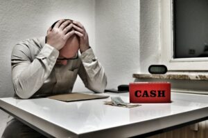 A person sits at a desk with their head in their hands, appearing stressed. On the desk are a red box labeled CASH, some money, and papers. The setting looks like an office or a small room.