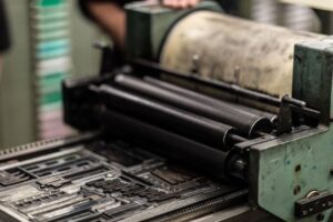 A close-up of a vintage printing press with ink rollers above a metal plate set with type, ready for printing. A persons arm is slightly visible in the background.