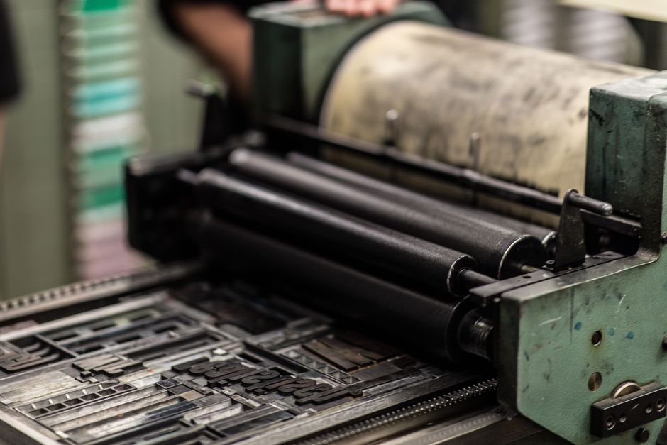 Printing Equipment Leasing: 2026 Guide For Print Shop Owners 1 A close-up of a vintage printing press with ink rollers above a metal plate set with type, ready for printing. A persons arm is slightly visible in the background.