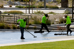 Three workers in bright green shirts and hats use wide squeegees to spread water across a paved surface in a landscaped outdoor area near grass and plants.