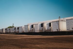 A row of white refrigerated semi-trailers is parked on a dirt lot in front of a white building under a clear blue sky.
