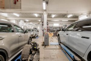 A car repair shop interior with several vehicles being serviced, tools and equipment scattered around, and white walls and ceiling.
