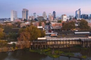 Aerial view of a city skyline at sunset with modern and historic buildings, trees with autumn foliage, and a river with a dock in the foreground.