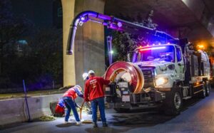 Two workers clean a street drain at night using a large vacuum truck with lights. One worker shovels debris while the other manages the hose attached to the truck under an overpass.
