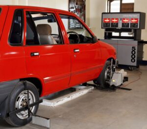 A red car undergoing a computer wheel alignment in a garage, with alignment equipment attached to the front wheels and a digital alignment system visible in the background.