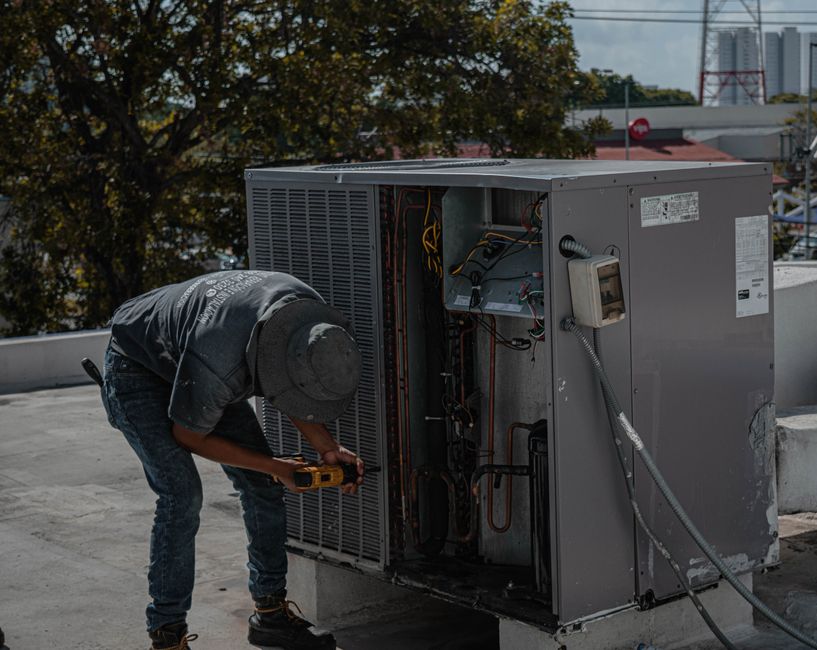 A person in a hat and gray shirt uses a power tool to repair or maintain a large hvac unit on a rooftop during the day.