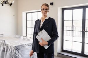 A woman in a dark suit holding a clipboard with documents stands in a bright room with large windows and a covered dining table. She is wearing glasses and looking at the camera.