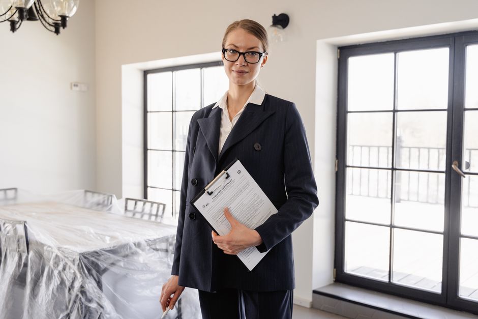 A woman in a dark suit holding a clipboard with documents stands in a bright room with large windows and a covered dining table. She is wearing glasses and looking at the camera.