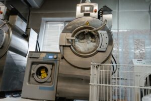 Two industrial washing machines filled with laundry are in a tiled laundry room. A metal laundry cart is visible in the foreground, and sunlight enters through a window behind the machines.