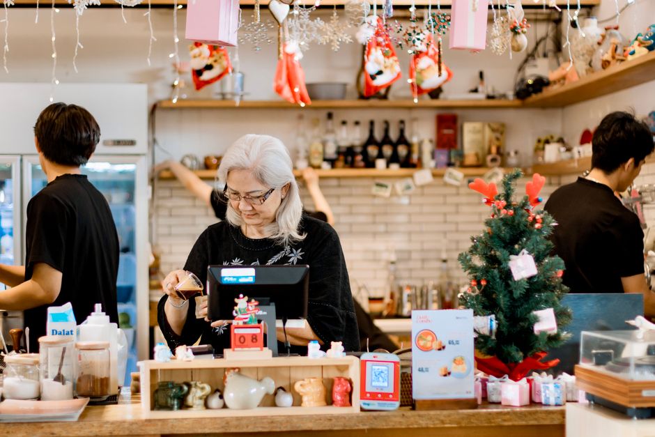 An older woman works at a cash register in a festive, christmas-themed cafe, with two people behind her preparing food and drinks. Holiday decorations and a small christmas tree are visible on the counter.