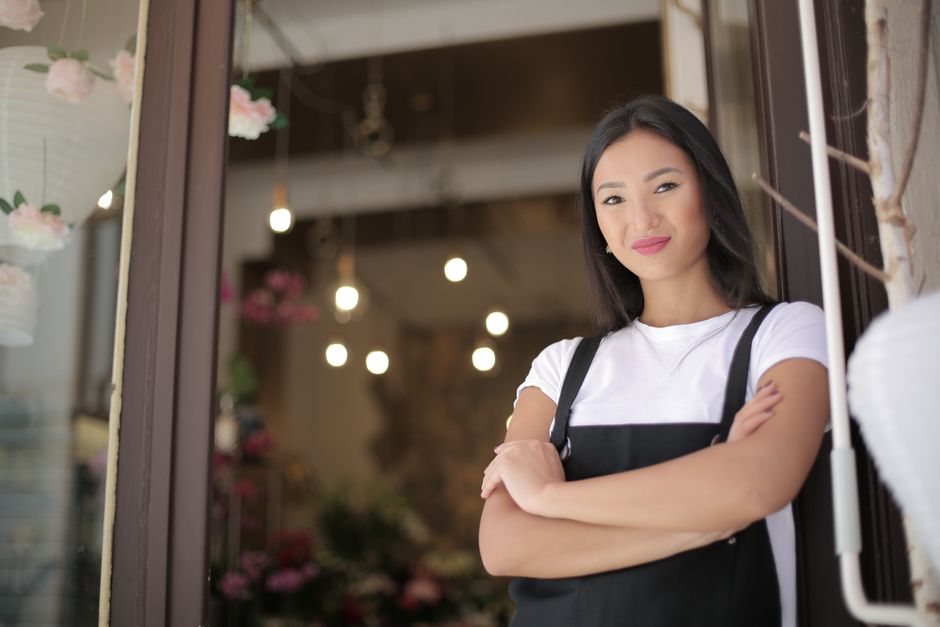 Woman in a black apron stands with arms crossed, smiling confidently at the entrance of a shop decorated with hanging lights and flowers.