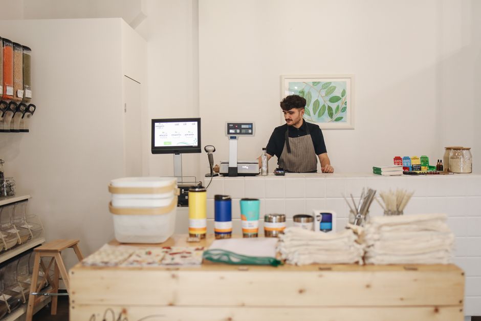 Man wearing an apron stands behind a shop counter with a computer. Reusable products and cloths are displayed in the foreground. Food dispensers are visible on the left wall. A leaf artwork hangs on the wall behind him.